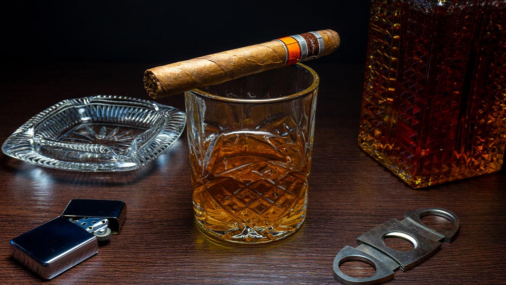 Trabuc pe un pahar de whisky langa o scrumiera, un cutter si o bricheta